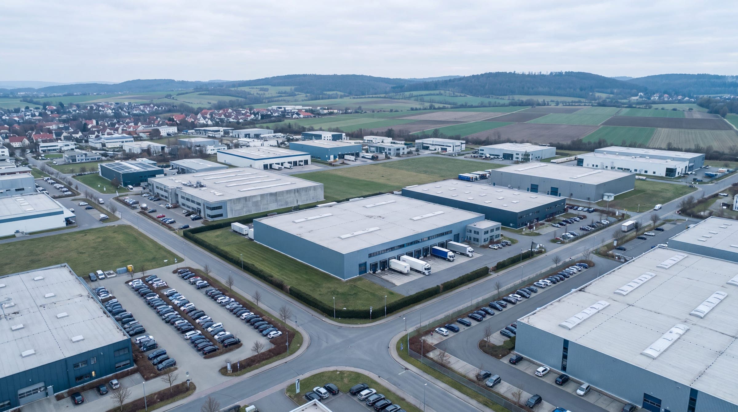 Luftaufnahme eines Gewerbegebiets am Ortsrand, dahinter Felder und bewaldete Hügel